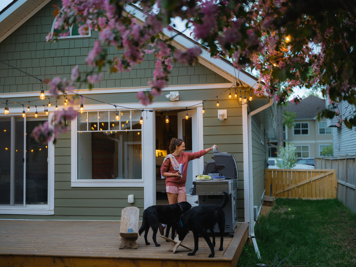 Woman grilling on a backyard deck with dogs, surrounded by string lights and pink flowering tree branches.